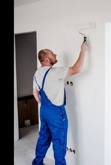Mężczyzna w niebieskim kombinezonie maluje ścianę wałkiem, widoczne puszki instalacyjne w ścianie, drzwi w tle.