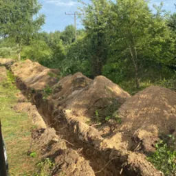 Długi, prosty rów w ziemi, z hałdami ziemi po bokach, częściowo porośnięty trawą i korzeniami, otoczony zielenią drzew i krzewów pod błękitnym niebem z lekkimi chmurami. Widoczny fragment maszyny...