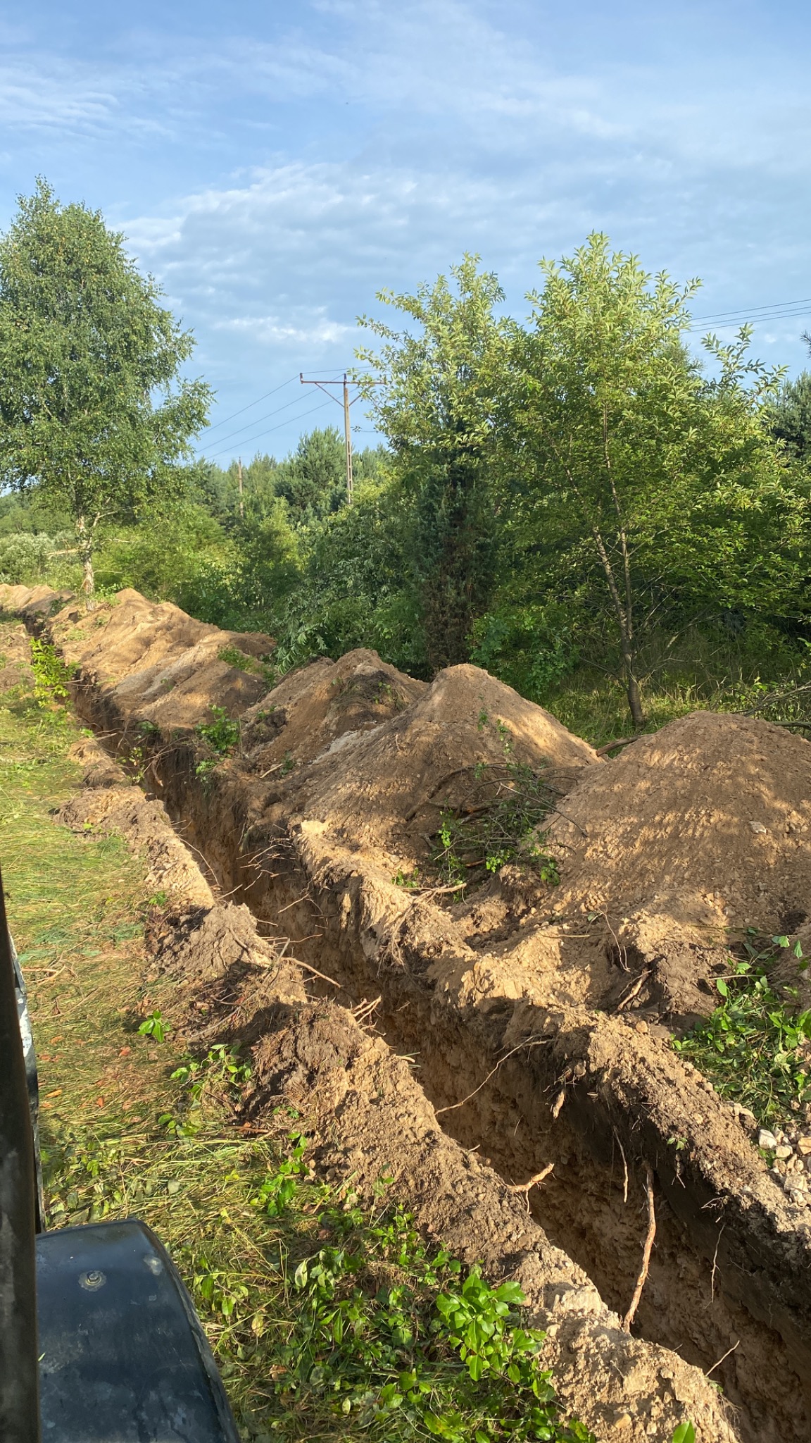 Długi, prosty rów w ziemi, z hałdami ziemi po bokach, częściowo porośnięty trawą i korzeniami, otoczony zielenią drzew i krzewów pod błękitnym niebem z lekkimi chmurami. Widoczny fragment maszyny...
