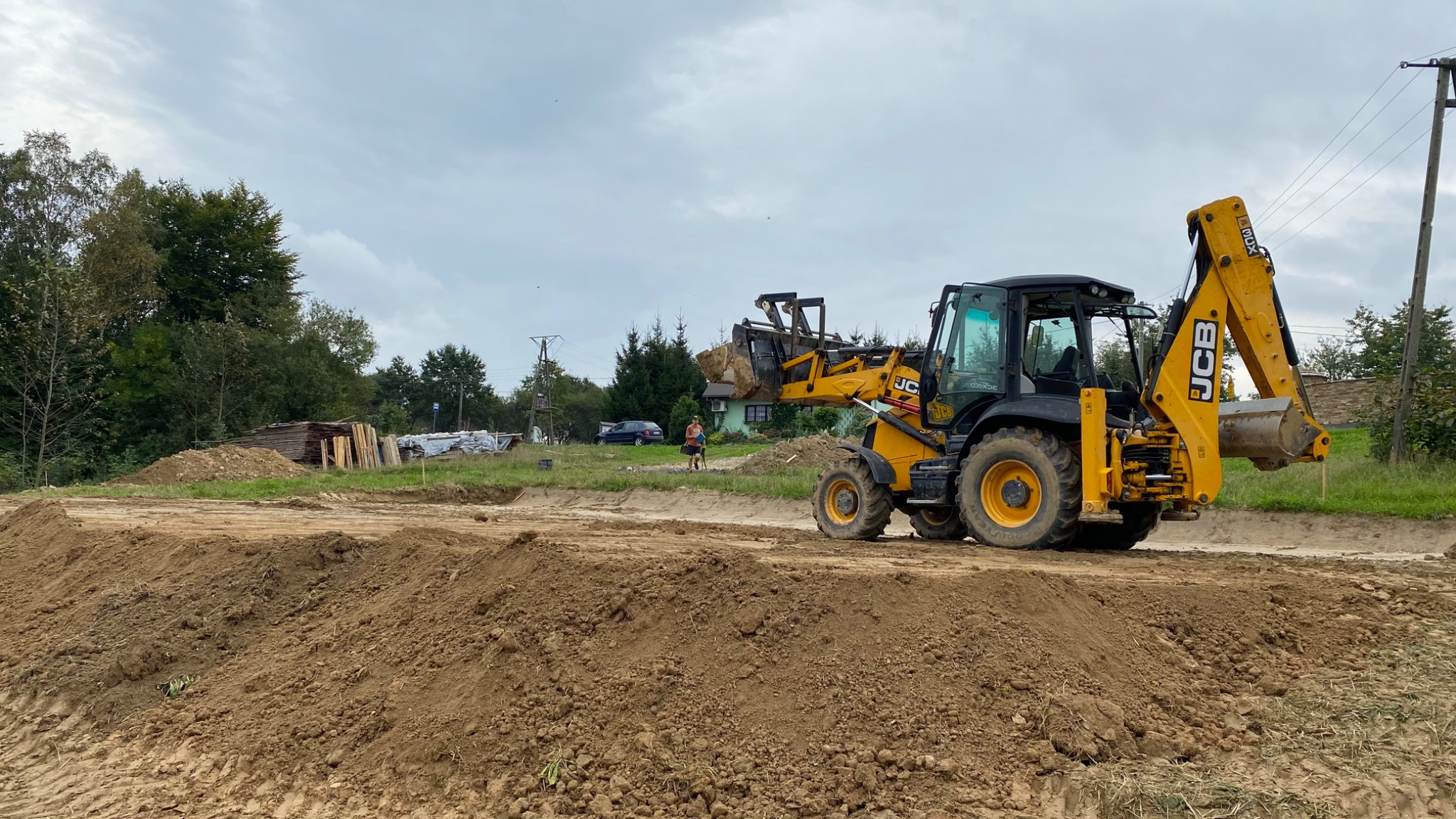 Żółto-czarna koparko-ładowarka JCB nabiera ziemię z wykopu, w tle widoczny dom i drzewa, pochmurne niebo.