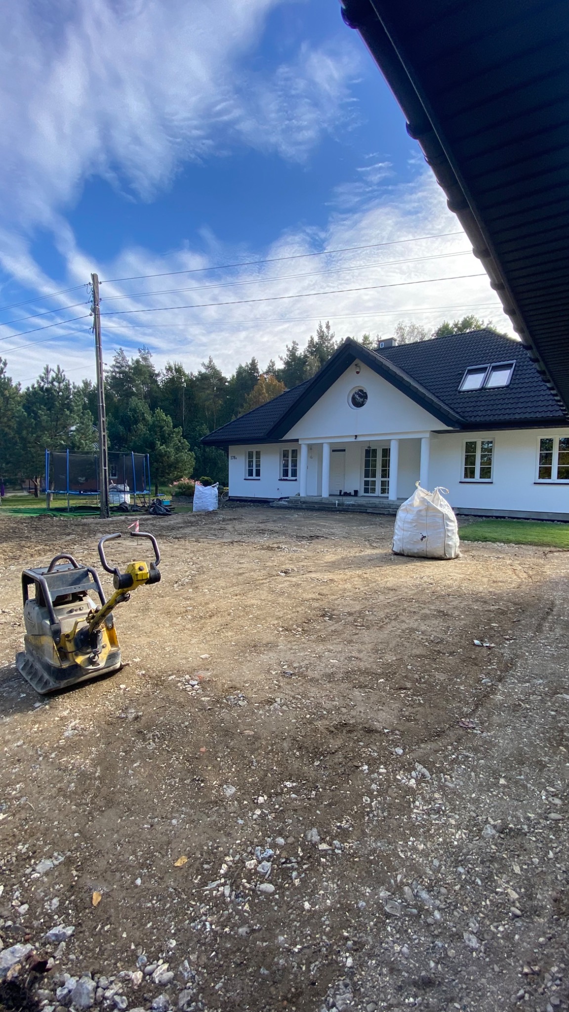 Żółto-czarna zagęszczarka wibrująca stoi na nierównym, kamienistym gruncie przed białym domem z czarnym dachem, worki z materiałem budowlanym w tle, błękitne niebo z chmurami.