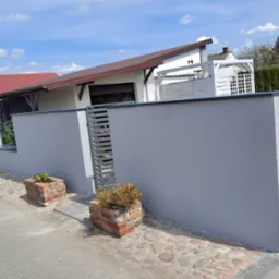 Świeżo otynkowany, gładki, szary mur posesji z metalowymi przęsłami, widok z poziomu ulicy, z kostką brukową i ceglanymi donicami.