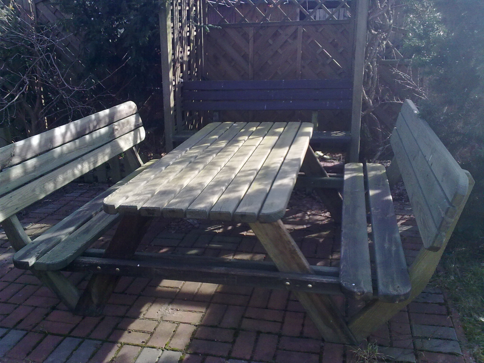 Drewniany stół piknikowy z ławkami, umieszczony na czerwonej kostce brukowej w ogrodzie, częściowo zacieniony, z widoczną drewnianą konstrukcją altany w tle.