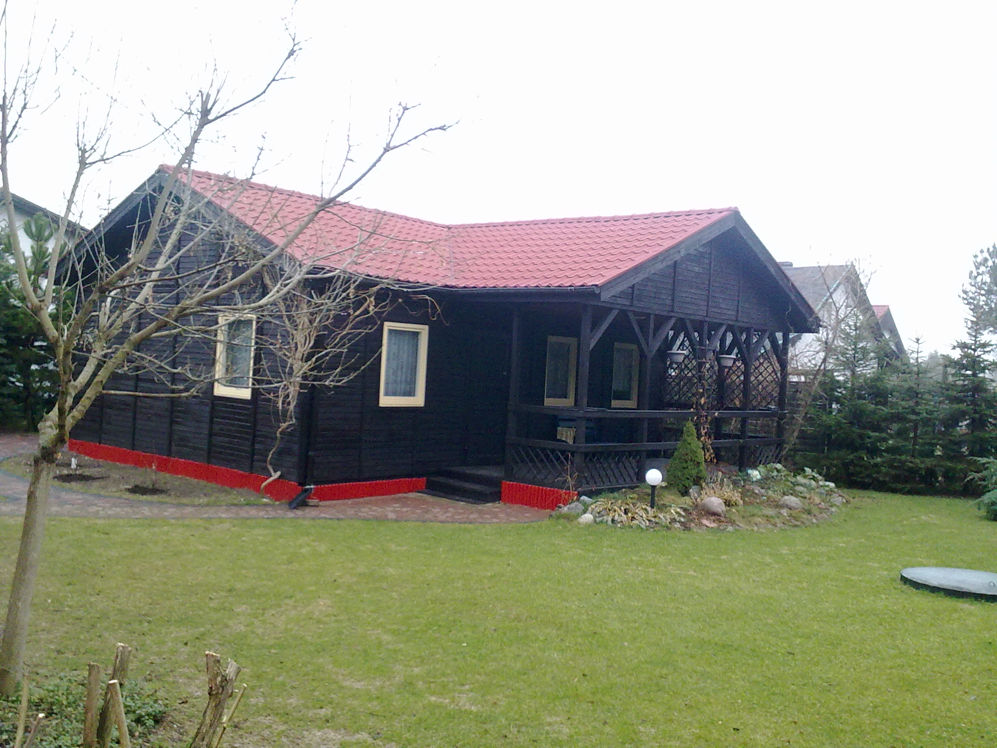 Czarna, drewniana altana z czerwonym dachem i podmurówką, widoczna z perspektywy ogrodu z trawnikiem i drzewami bez liści.