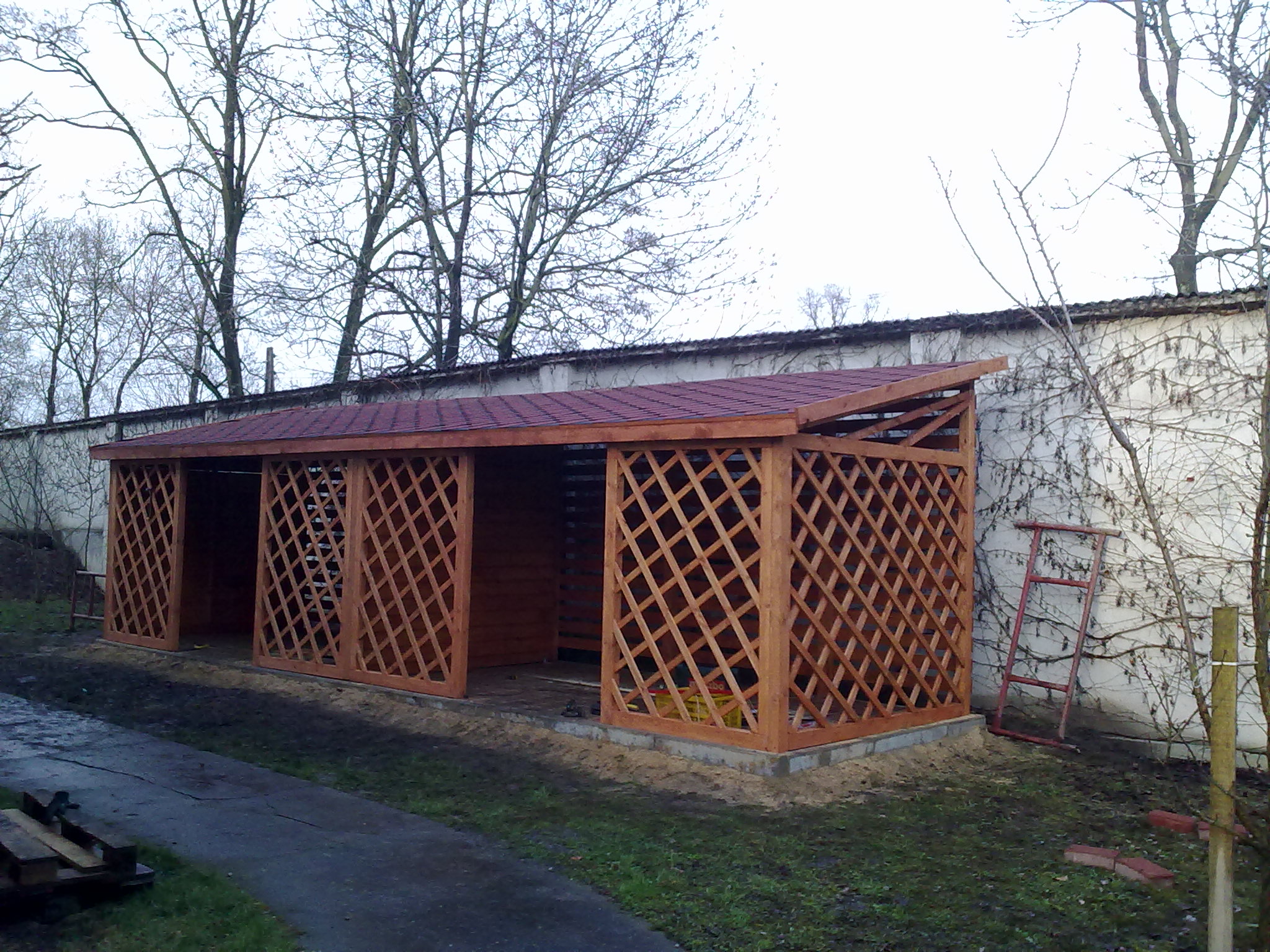 Drewniana altana z kratownicowymi ścianami i czerwonym dachem, częściowo ukończona, na tle białego muru z pnączami i drzew bez liści, z drabiną opartą o ścianę po prawej stronie.
