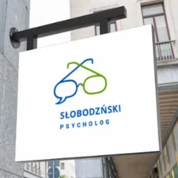 Szyld reklamowy z logo w kształcie okularów i dymku dialogowego, w kolorach niebieskim i zielonym, z nazwą 'SŁOBODZIŃSKI PSYCHOLOG' na tle budynku.