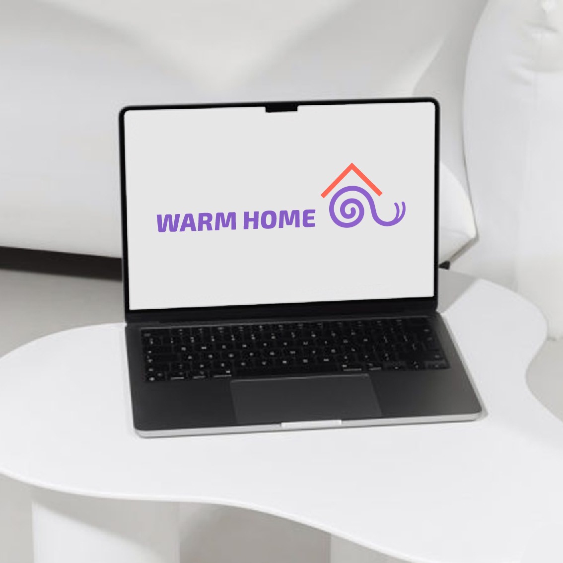 Laptop wyświetlający logo 'Warm Home' z symbolem domu i spiralą na białym tle, umieszczony na białym stoliku w minimalistycznym wnętrzu.