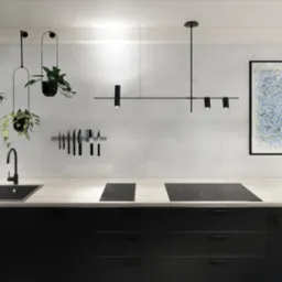 Nowoczesna kuchnia z czarnymi szafkami, jasnym blatem, czarnym zlewozmywakiem, indukcyjną płytą grzewczą, wiszącymi roślinami w czarnych doniczkach, oświetleniem punktowym i abstrakcyjnym obrazem...