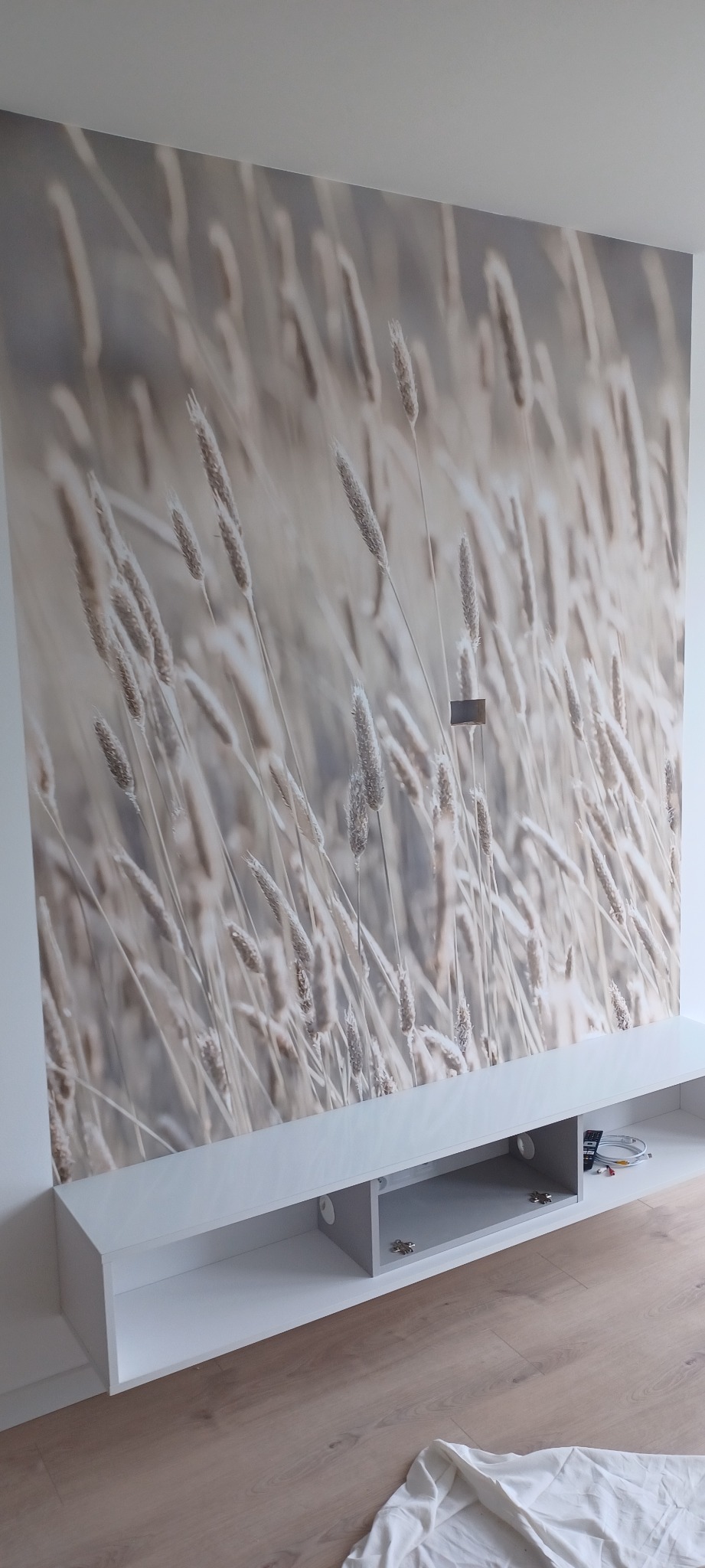 Ściana z fototapetą przedstawiającą motyw trawy, pod nią biała szafka RTV z otwartymi półkami. Na podłodze leży biała tkanina.
