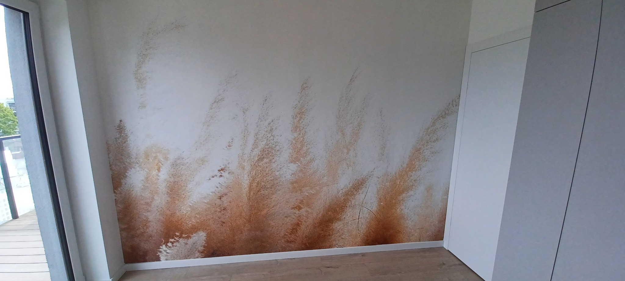 Ściana w pokoju z fototapetą przedstawiającą pampasową trawę, widoczna część okna balkonowego i biała szafa wnękowa.