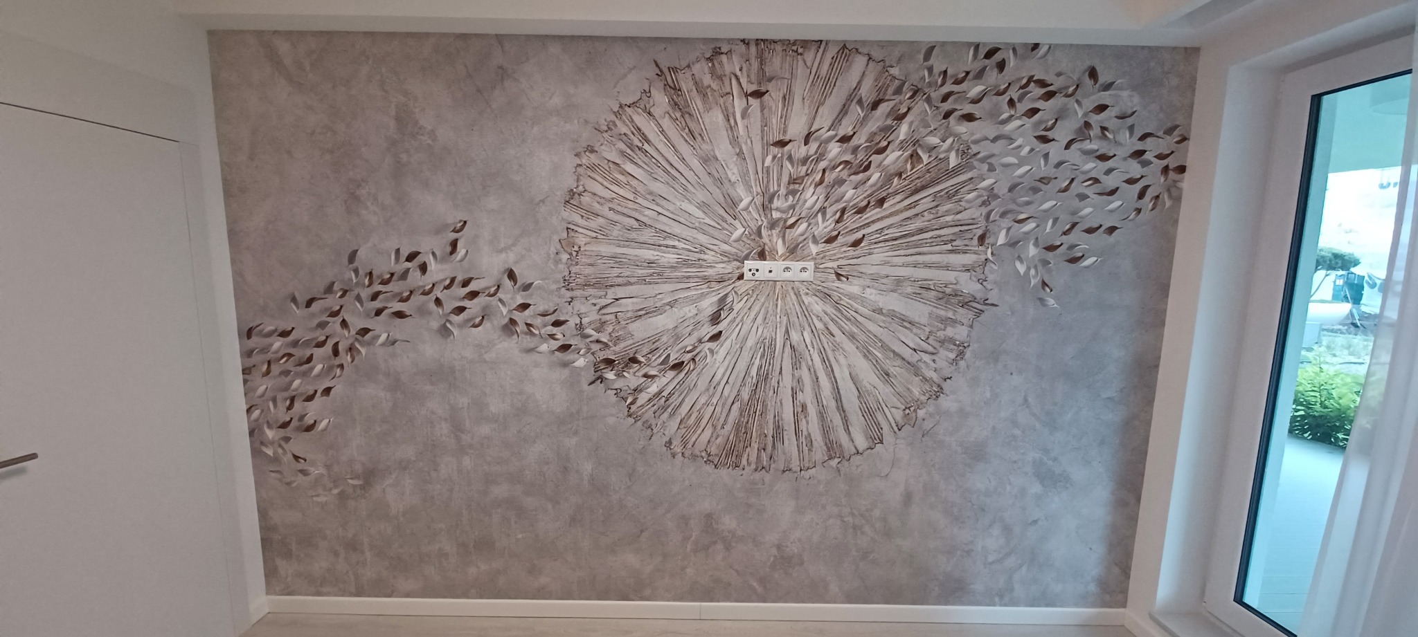 Ściana z dekoracyjną fakturą w odcieniach szarości, zdobiona wypukłym motywem rozchodzących się promieni i odrywających się listków. Gniazdka elektryczne wkomponowane w centrum kompozycji.