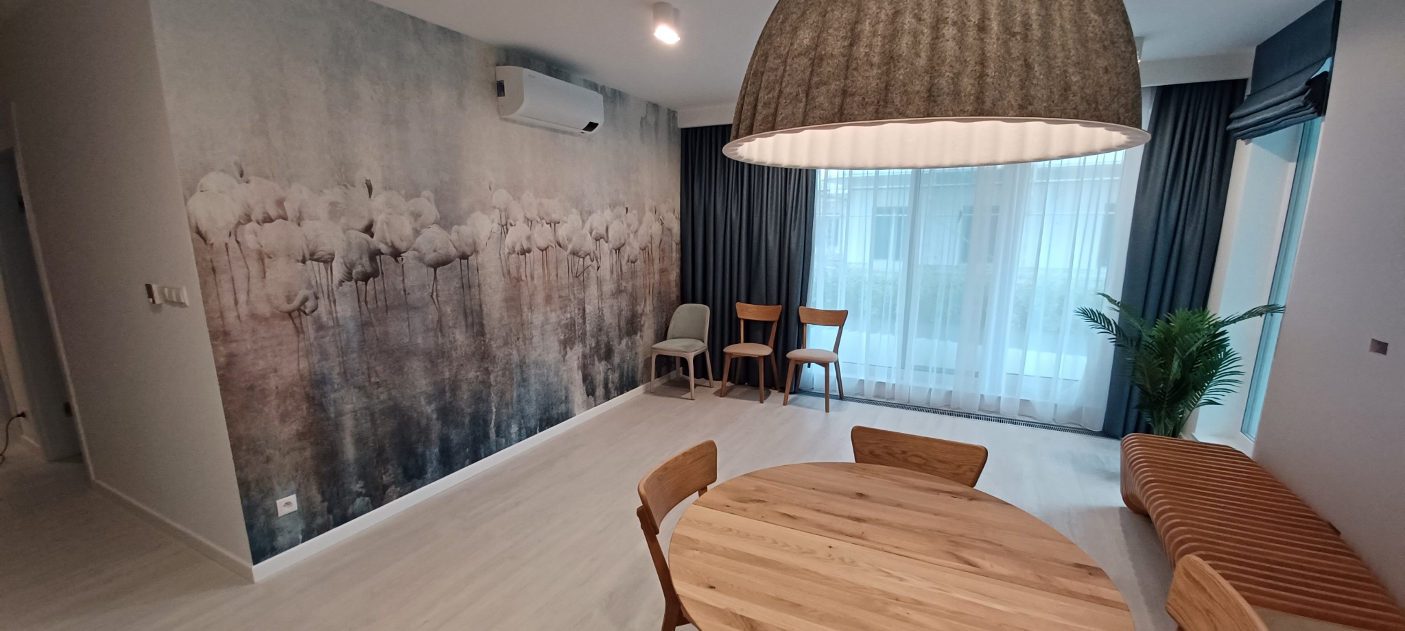 Jasne wnętrze jadalni z okrągłym drewnianym stołem, krzesłami i tapetą z motywem flamingów, oświetlone wiszącą lampą w kształcie kapelusza.