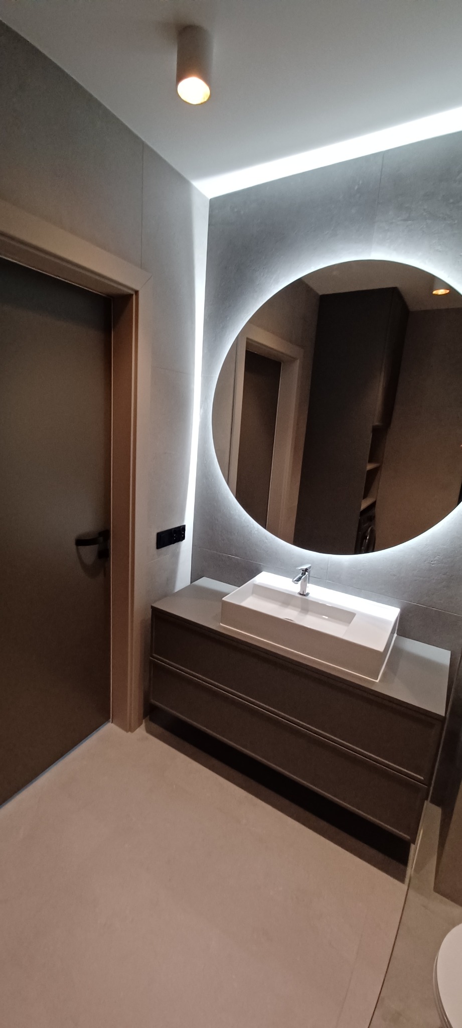Nowoczesna łazienka z umywalką nablatową, szafką z szufladami i okrągłym lustrem z podświetleniem LED, widok na drzwi i minimalistyczne oświetlenie sufitowe.