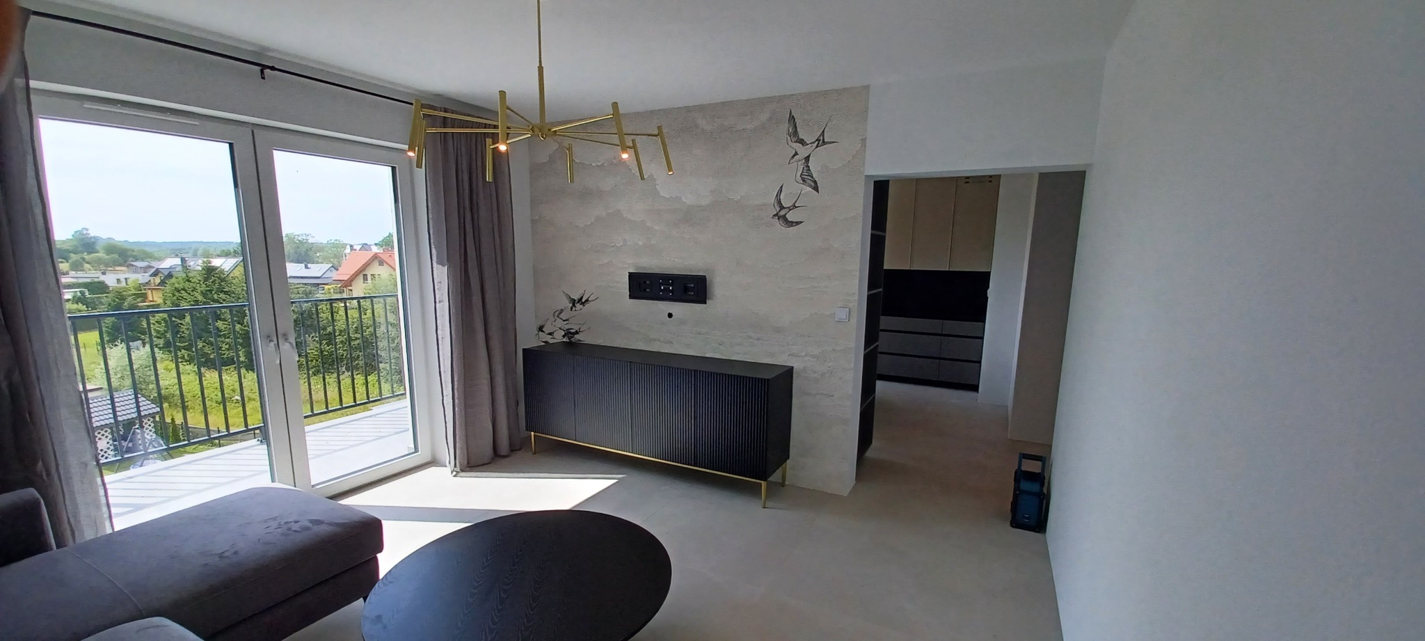 Nowoczesny salon z czarną komodą na złotych nóżkach, abstrakcyjną tapetą z motywem ptaków, złotą lampą wiszącą i wyjściem na balkon z widokiem na zieleń.