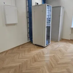 Serwerownia z szafami rack, korytkami kablowymi pod sufitem i pionowym prowadzeniem niebieskich kabli w biurowym wnętrzu z parkietem w jodełkę.