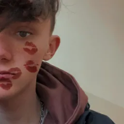 Młody mężczyzna z pomalowanymi ustami na policzku, ubrany w bluzę z kapturem i srebrny łańcuszek.