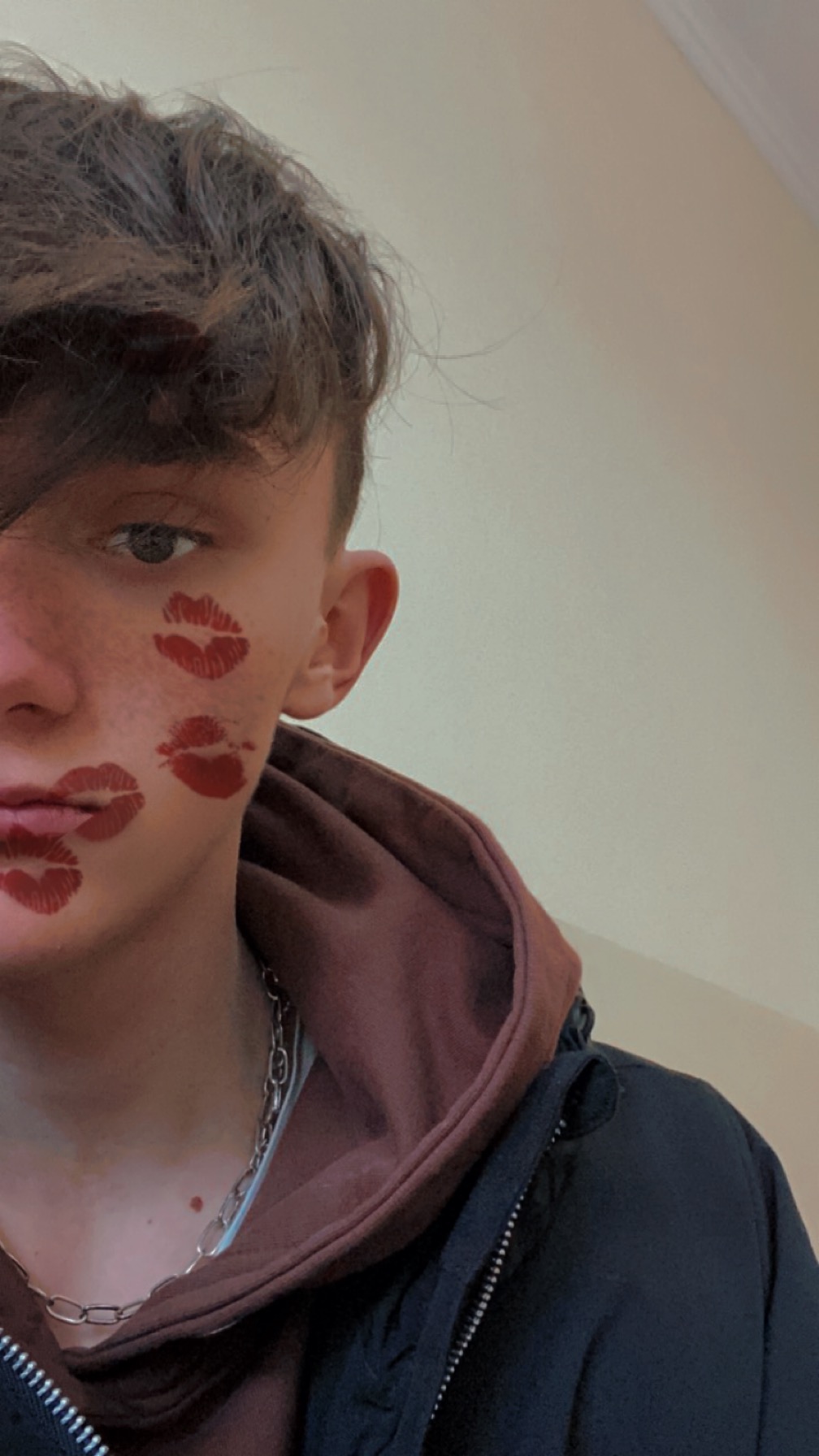 Młody mężczyzna z pomalowanymi ustami na policzku, ubrany w bluzę z kapturem i srebrny łańcuszek.