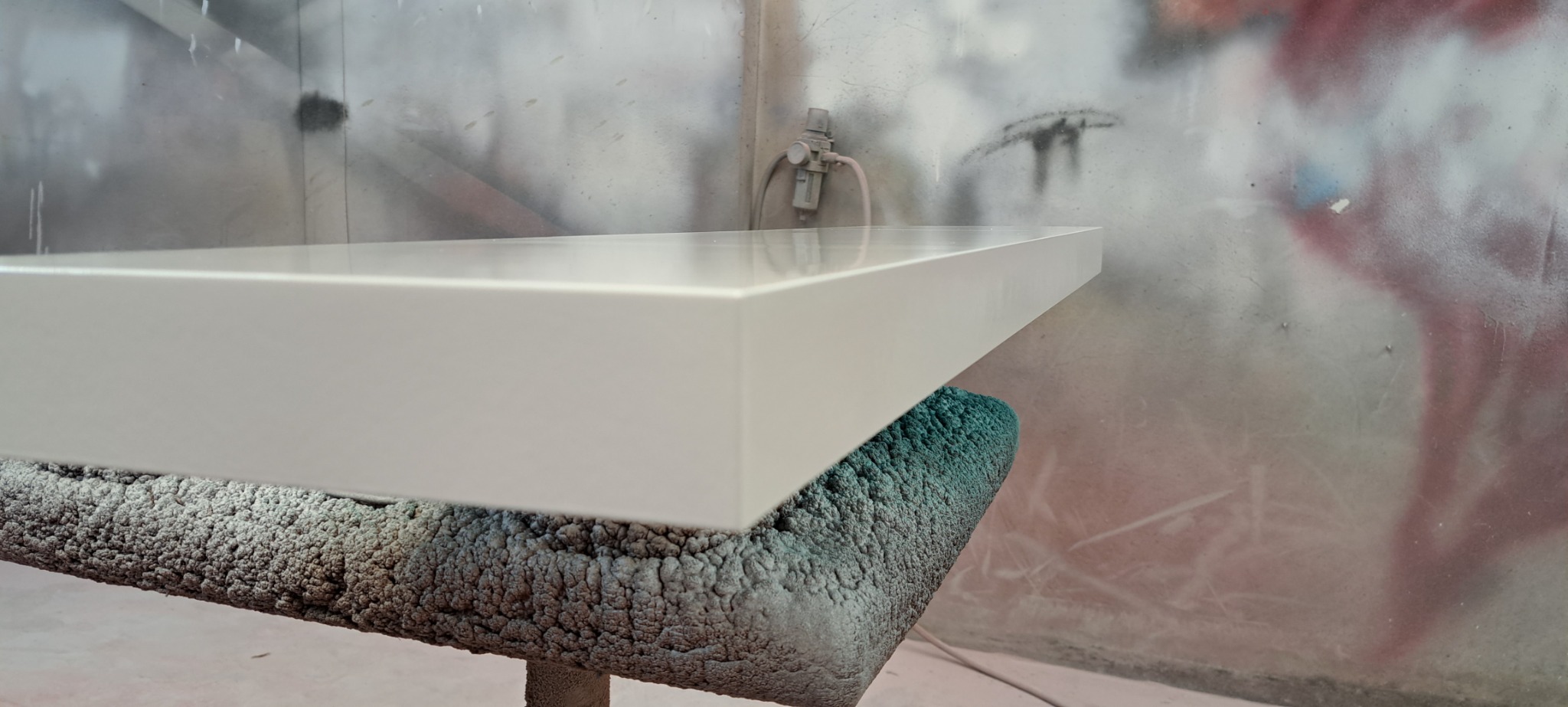 Lakierowany na biało blat drewniany na stojaku w lakierni, widoczny pistolet lakierniczy w tle.