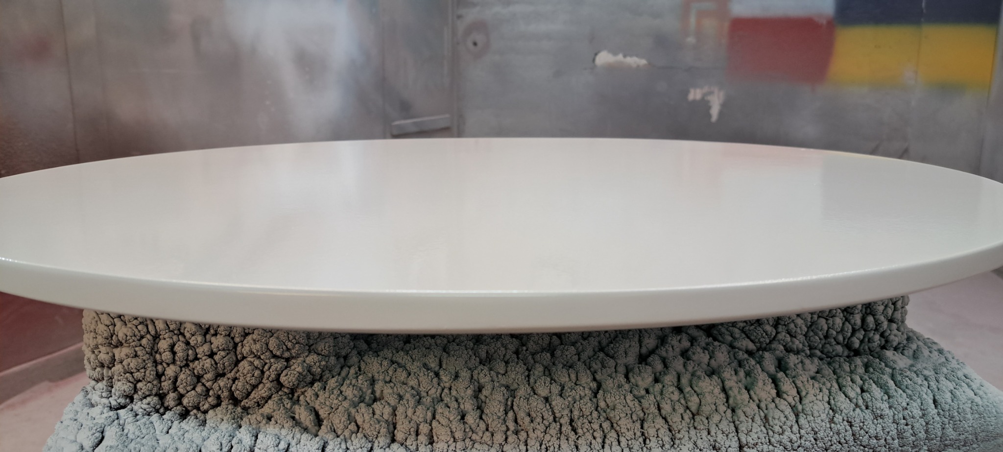 Biały, okrągły blat poddawany obróbce lakierniczej, wsparty na chropowatej, szarej podstawie w kabinie lakierniczej.