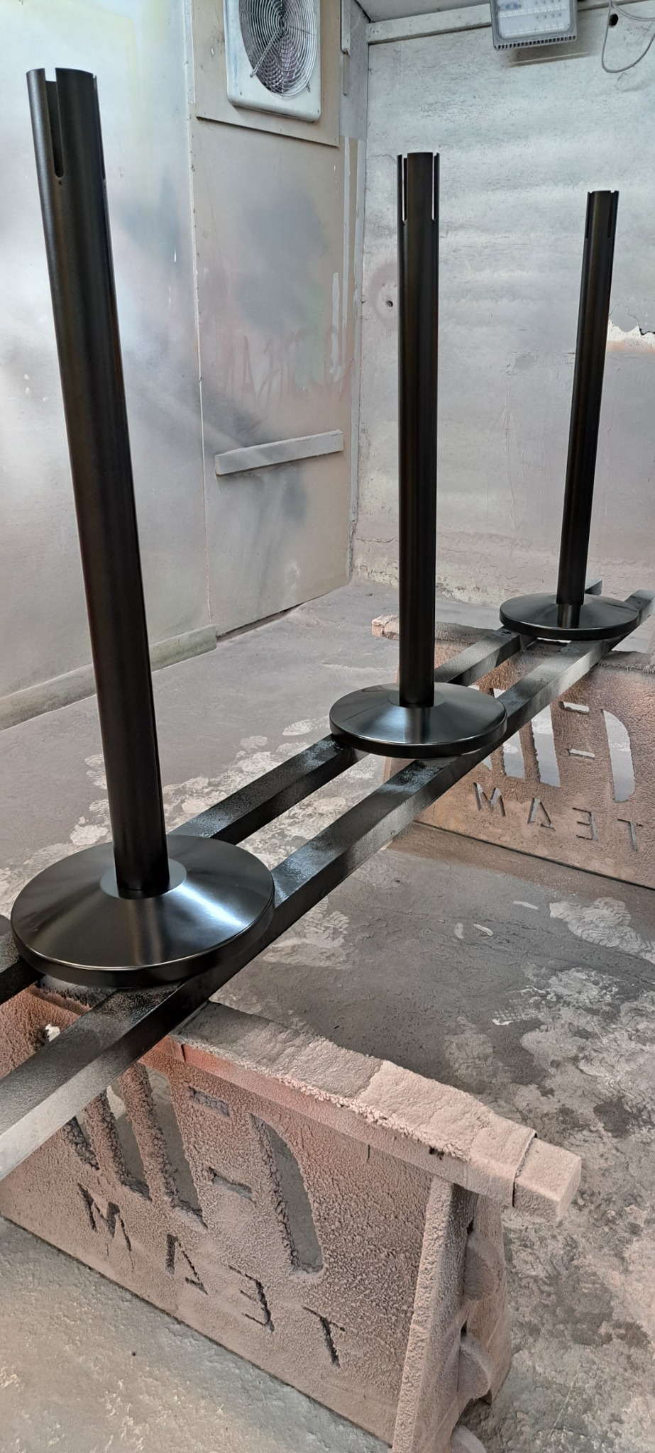 Trzy czarne, metalowe nogi stołowe z okrągłymi podstawami, ustawione na metalowej konstrukcji w warsztacie, gotowe do dalszej obróbki lub montażu.