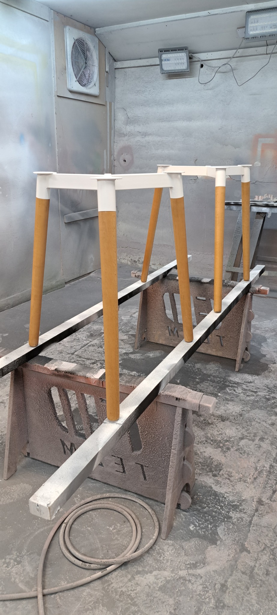 Stojaki pod blaty z drewnianymi nogami w trakcie lakierowania w lakierni, widoczne detale konstrukcji i niedoskonałości podłoża.