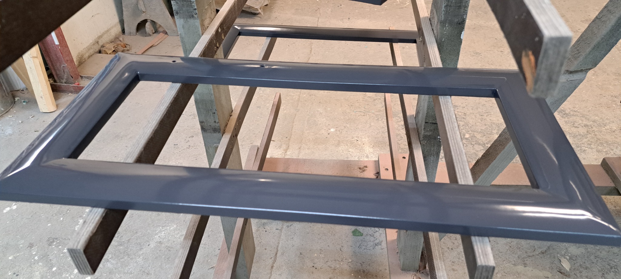 Szare, lakierowane ramy drzwiowe lub meblowe, leżące na drewnianych kozłach w warsztacie, widoczne otwory montażowe.