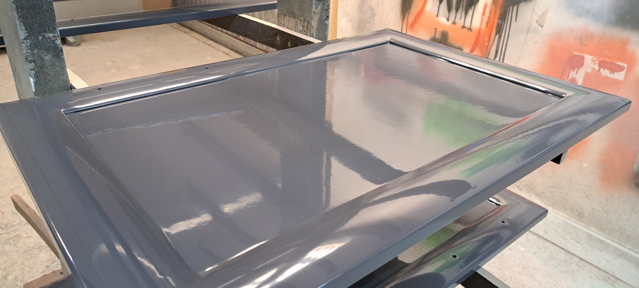 Szare, lakierowane fronty meblowe z widocznymi otworami montażowymi, ułożone na metalowym stojaku w warsztacie.