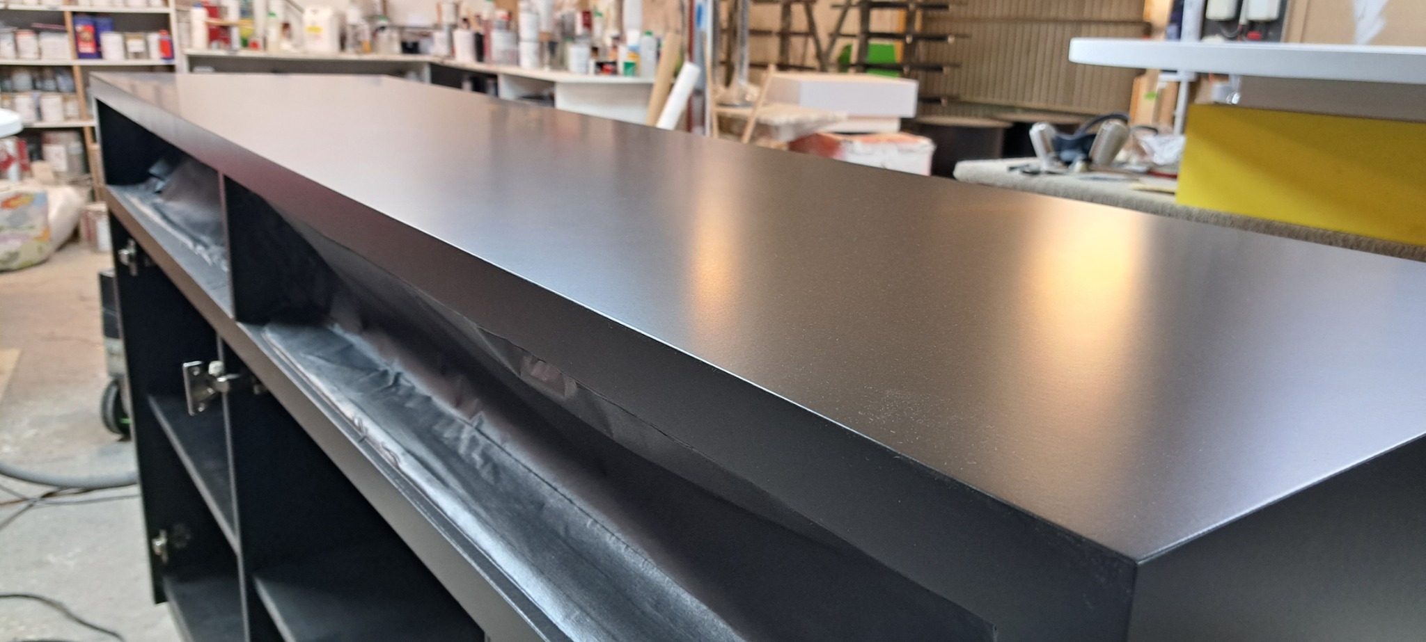 Ciemny, lakierowany blat z widocznymi półkami i drzwiczkami na metalowych zawiasach, sfotografowany w warsztacie stolarskim.