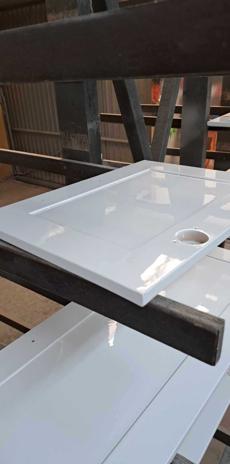 Białe, błyszczące fronty meblowe z frezowanymi krawędziami, suszone na drewnianym regale w warsztacie.