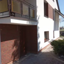 Dwukondygnacyjny dom z białą elewacją, fragmentem ściany wykończonym czerwoną cegłą klinkierową i balkonem z ozdobną balustradą, widziany pod kątem z poziomu chodnika.