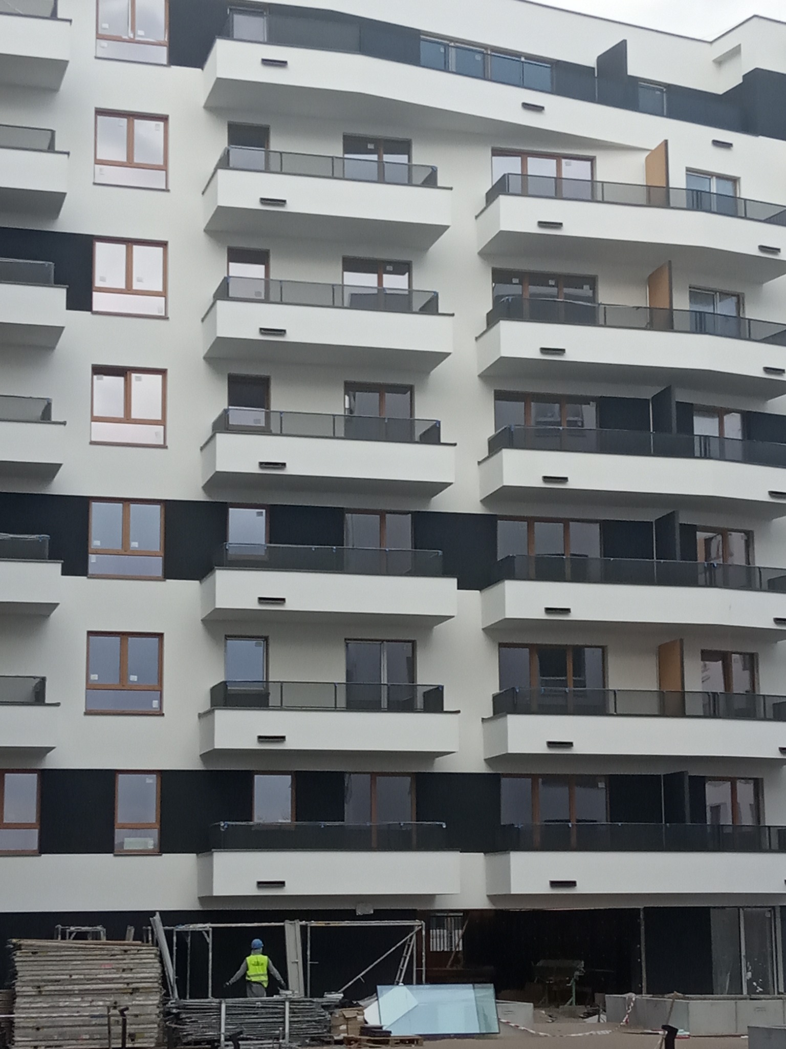 Wielopiętrowy budynek z balkonami, częściowo ocieplony, z widocznym pracownikiem w kamizelce odblaskowej i niebieskim kasku oraz materiałami budowlanymi na pierwszym planie.
