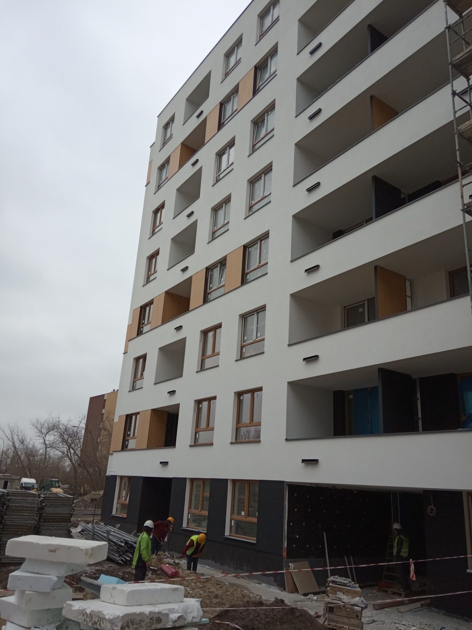 Wielopiętrowy budynek mieszkalny w trakcie prac wykończeniowych elewacji, widoczne rusztowania i pracownicy w odblaskowych kamizelkach na placu budowy.
