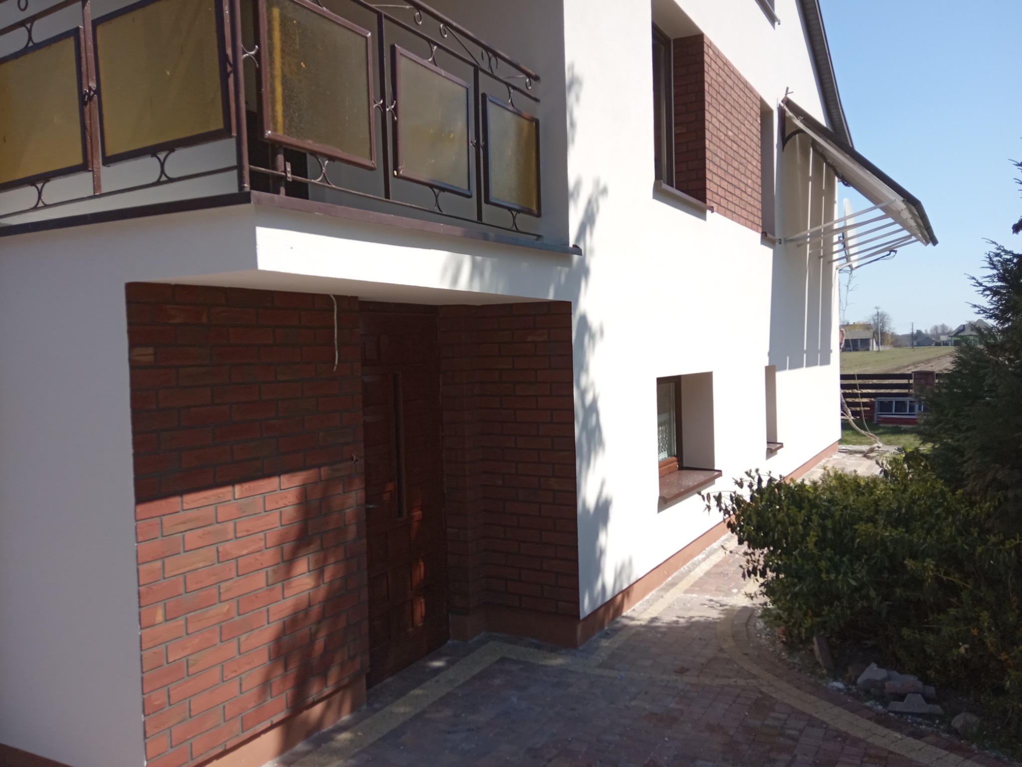 Dwukondygnacyjny dom z białą elewacją, fragmentem ściany wykończonym czerwoną cegłą klinkierową i balkonem z ozdobną balustradą, widziany pod kątem z poziomu chodnika.