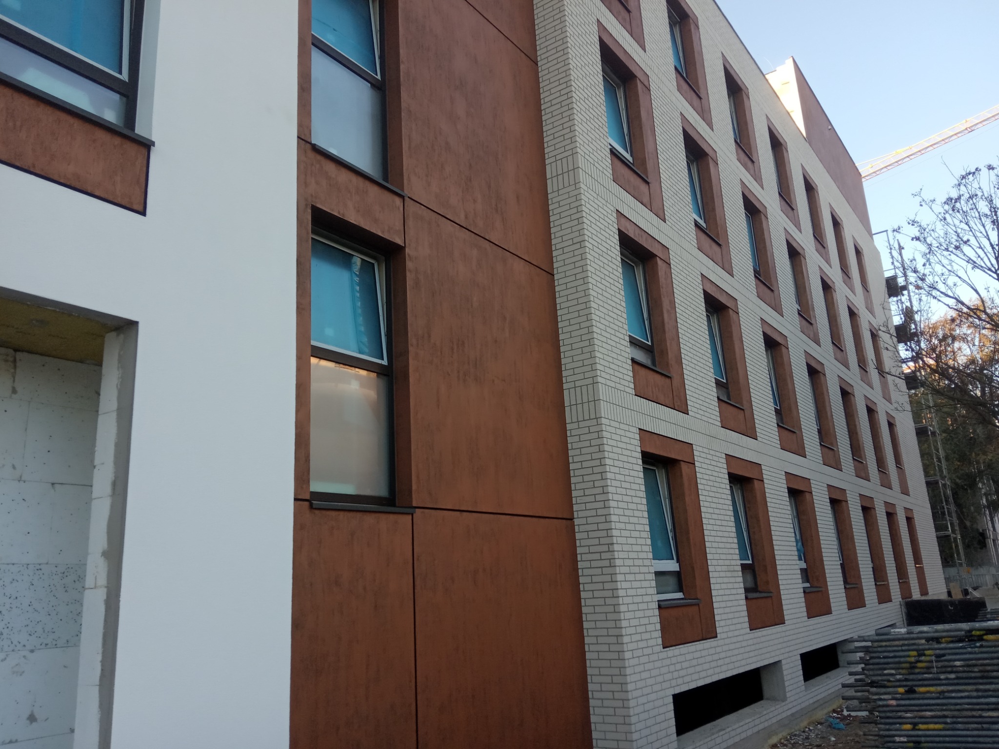 Wielopiętrowy budynek z fragmentami elewacji wykończonymi panelami w odcieniach brązu i jasną cegłą, okna zabezpieczone folią, widoczny żuraw budowlany w tle.