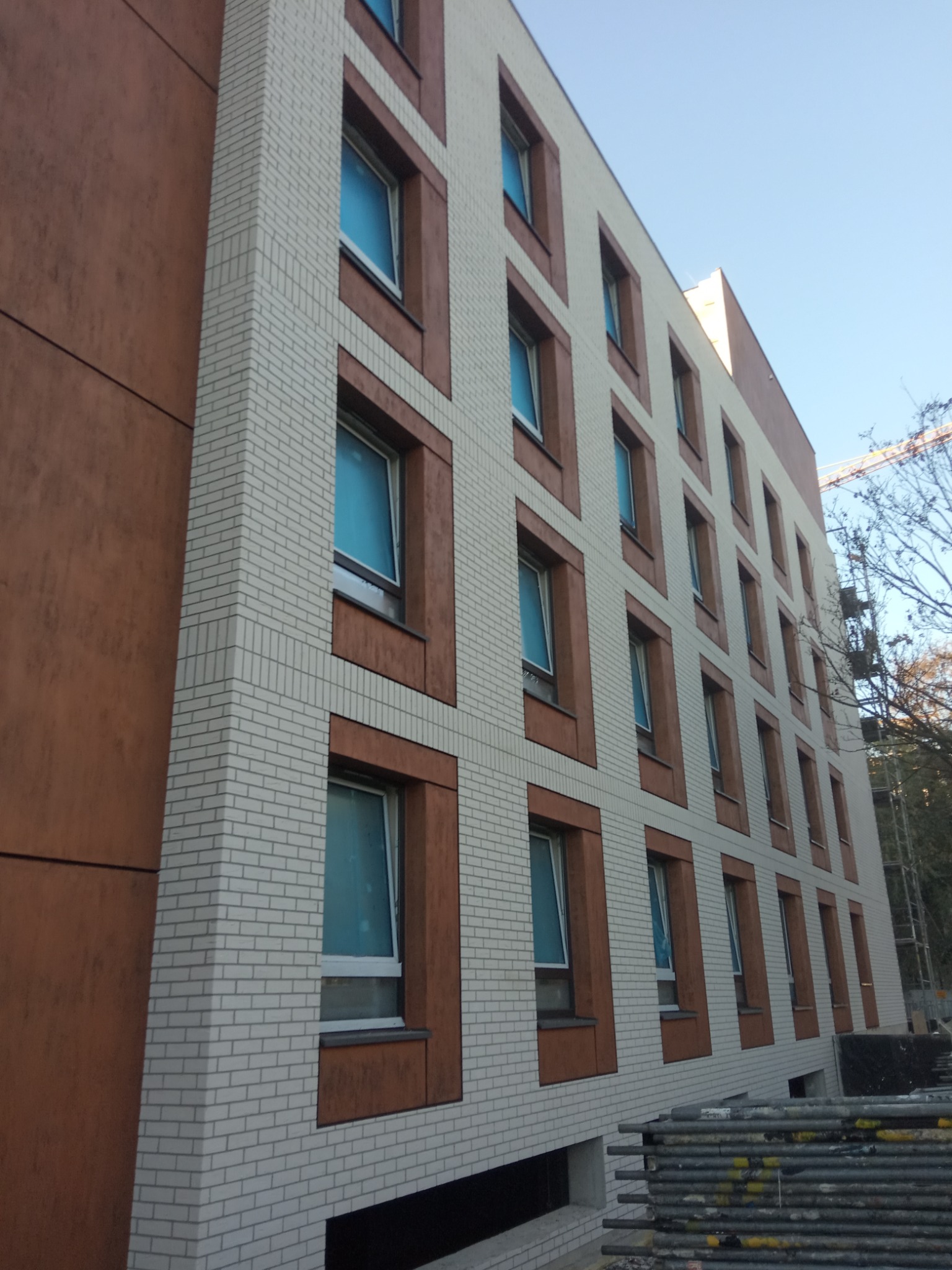 Wielopiętrowy budynek z jasnej cegły i brązowych paneli elewacyjnych, widok pod kątem z widocznym żurawiem budowlanym w tle.