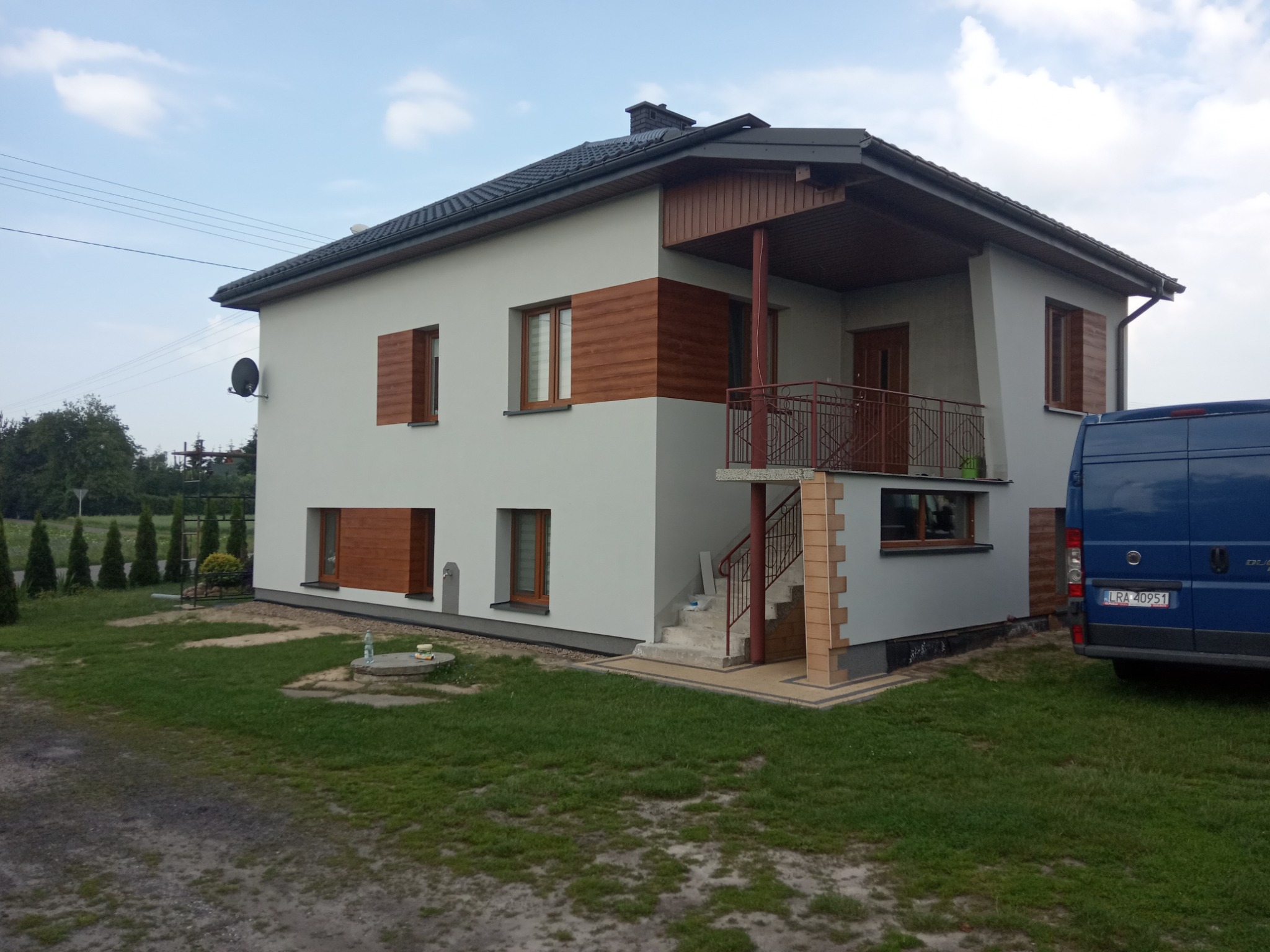 Dwukondygnacyjny dom jednorodzinny z szarą elewacją i drewnianymi elementami dekoracyjnymi, w tle błękitny samochód dostawczy.