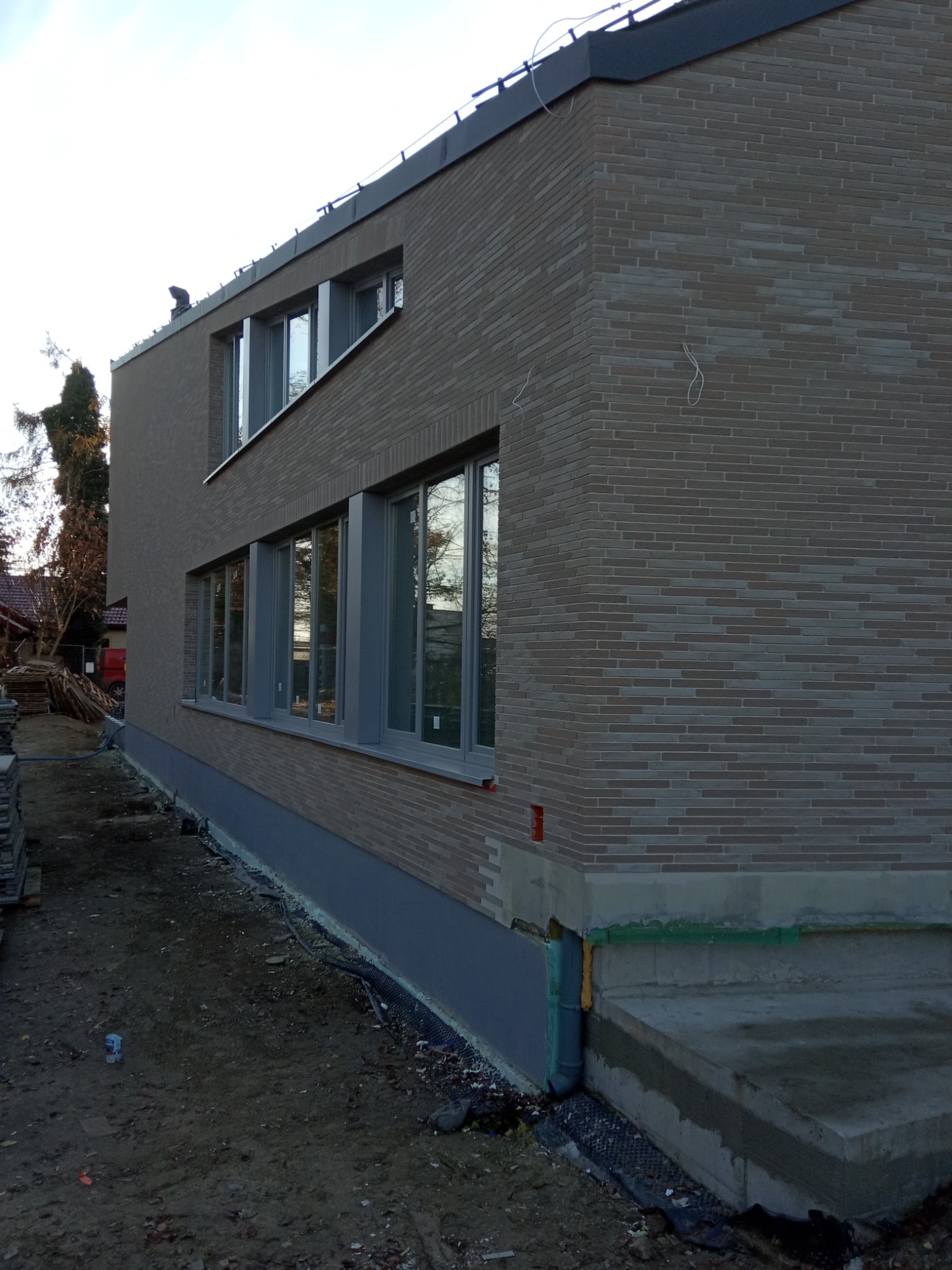 Nowoczesny budynek z elewacją z cegły klinkierowej w trakcie wykańczania, widoczne przygotowane fundamenty i okna z szarymi ramami.