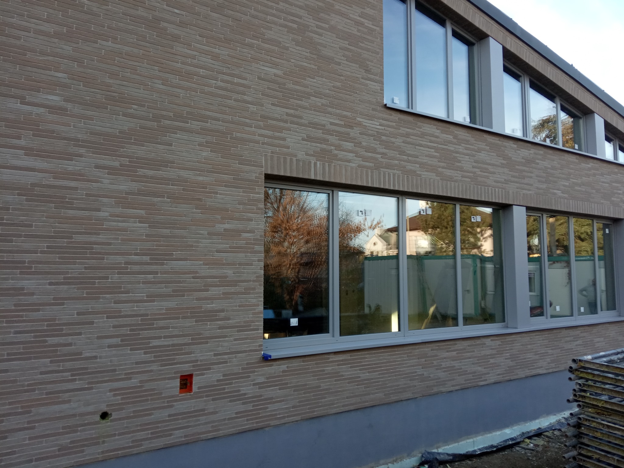 Elewacja budynku wykończona cegłą klinkierową w odcieniach beżu i brązu, widoczne okna z refleksami otoczenia, fragment rusztowania w prawym dolnym rogu.