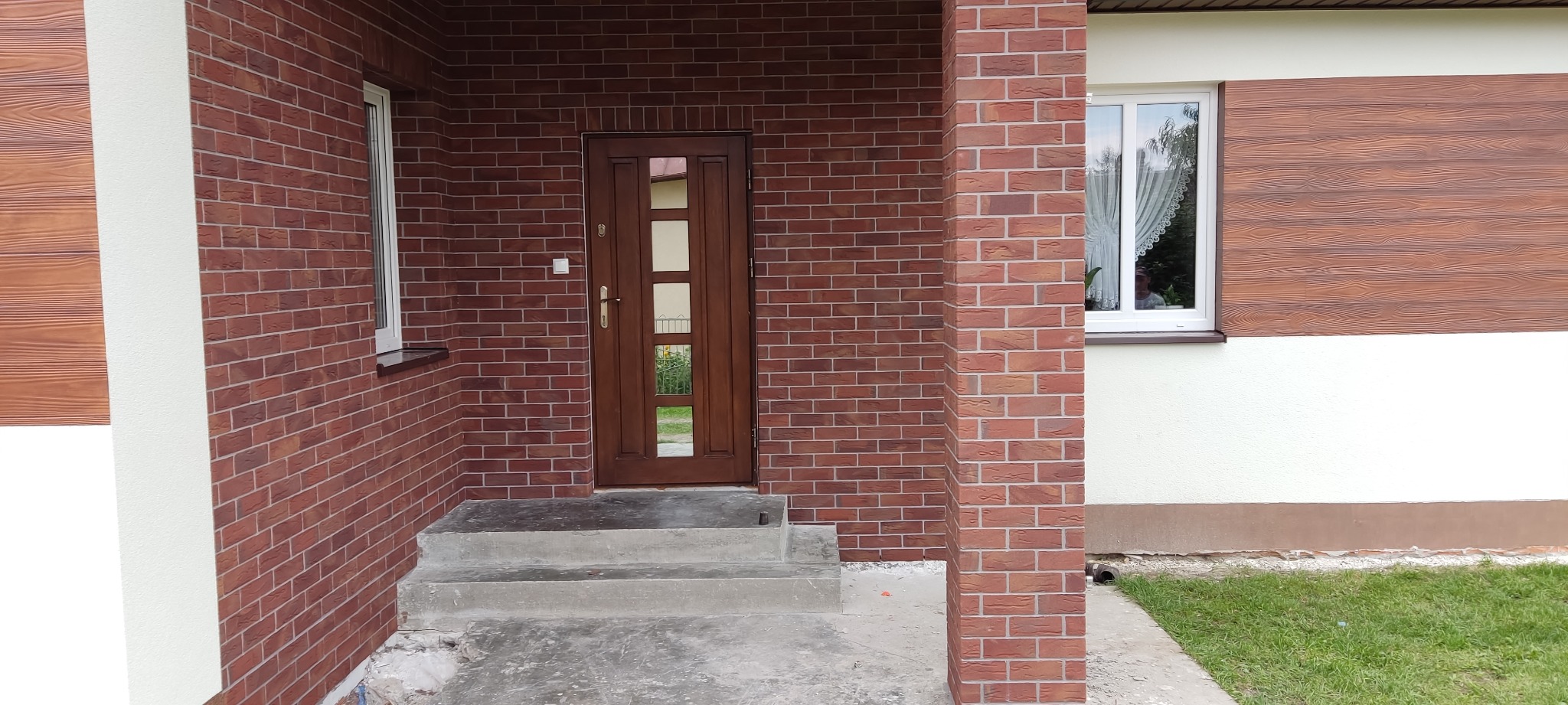 Wykończona elewacja domu z cegły klinkierowej, brązowe drewniane drzwi wejściowe z przeszkleniami, białe okna i imitacja drewnianej okładziny elewacyjnej.