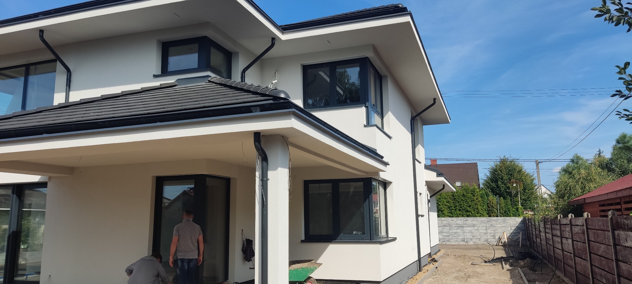 Nowoczesny dom jednorodzinny z jasną elewacją, ciemnymi oknami i dachem, widoczne prace wykończeniowe wokół budynku.