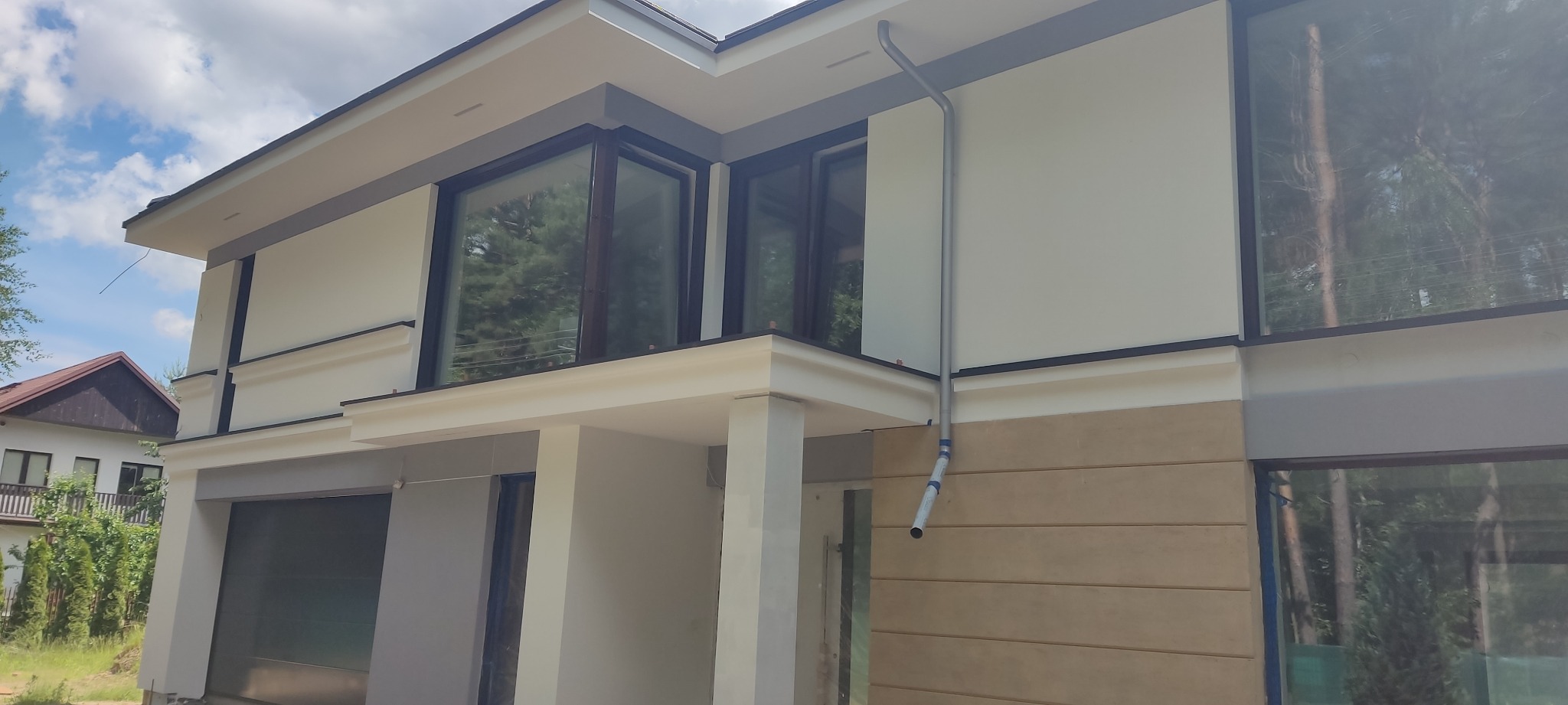 Nowoczesny dom jednorodzinny z jasną elewacją, dużymi oknami z ciemnymi ramami i widoczną rynną spustową, częściowo otynkowany, z elementami dekoracyjnymi.