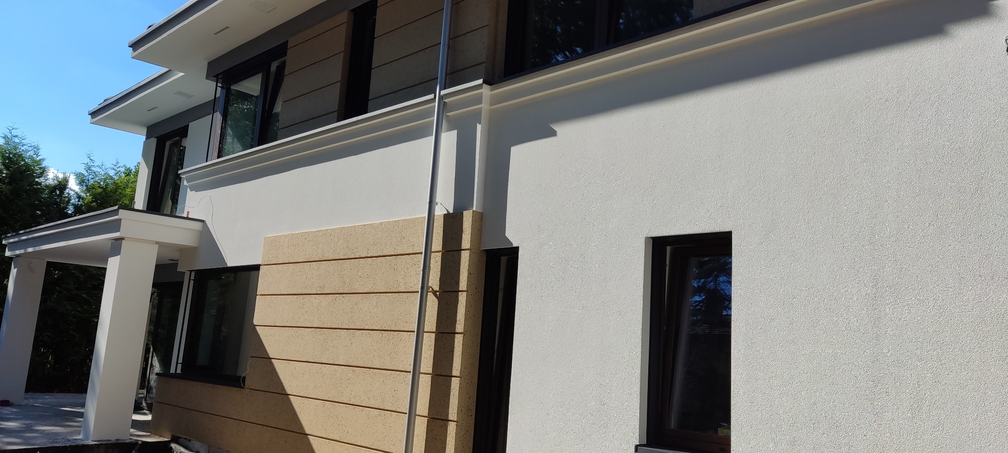 Fragment elewacji domu z widoczną strukturą tynku baranka w kolorze białym oraz elementem dekoracyjnym z paneli imitujących drewno, pionowa metalowa rura przy ścianie.