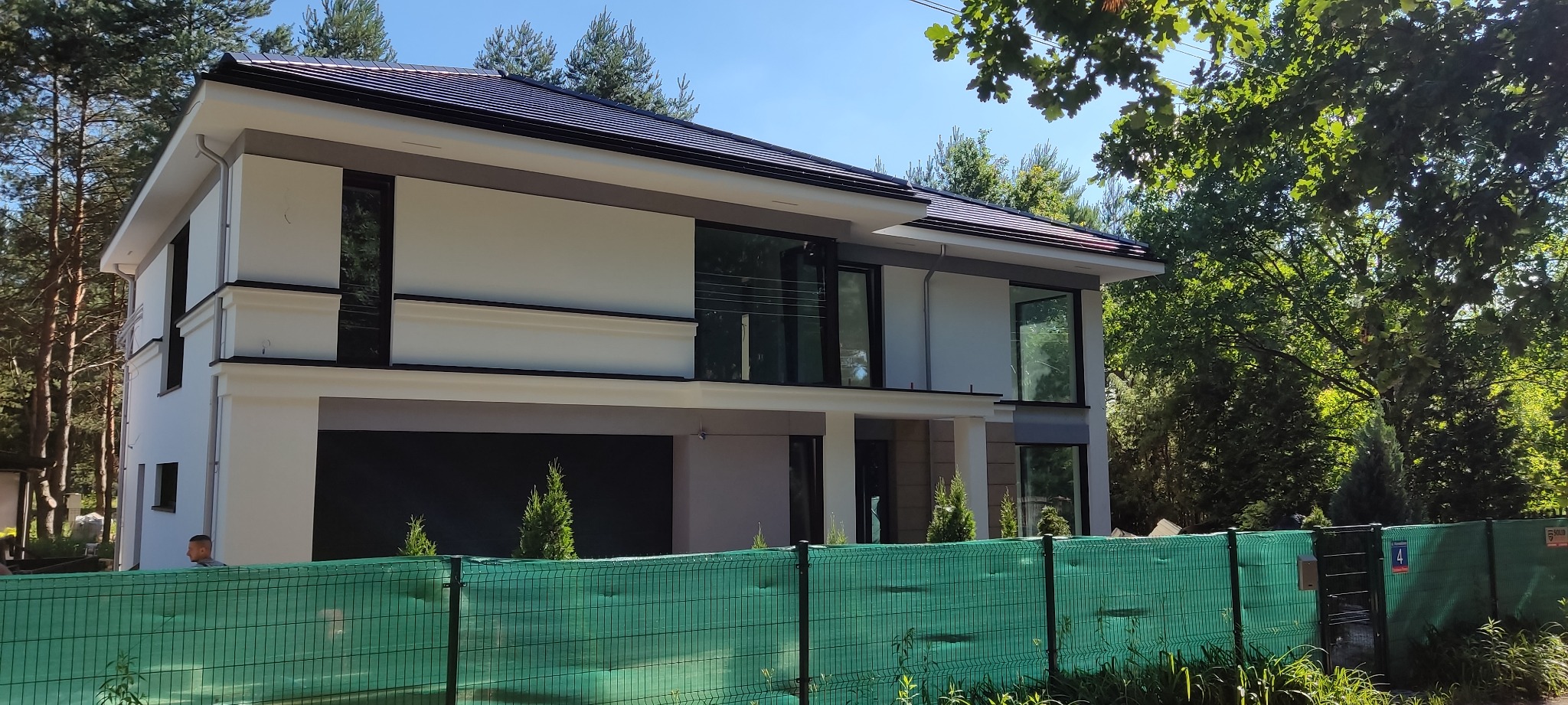 Nowoczesny dom jednorodzinny z białą elewacją, dużymi oknami i ciemną dachówką, widoczny za zielonym ogrodzeniem budowlanym.