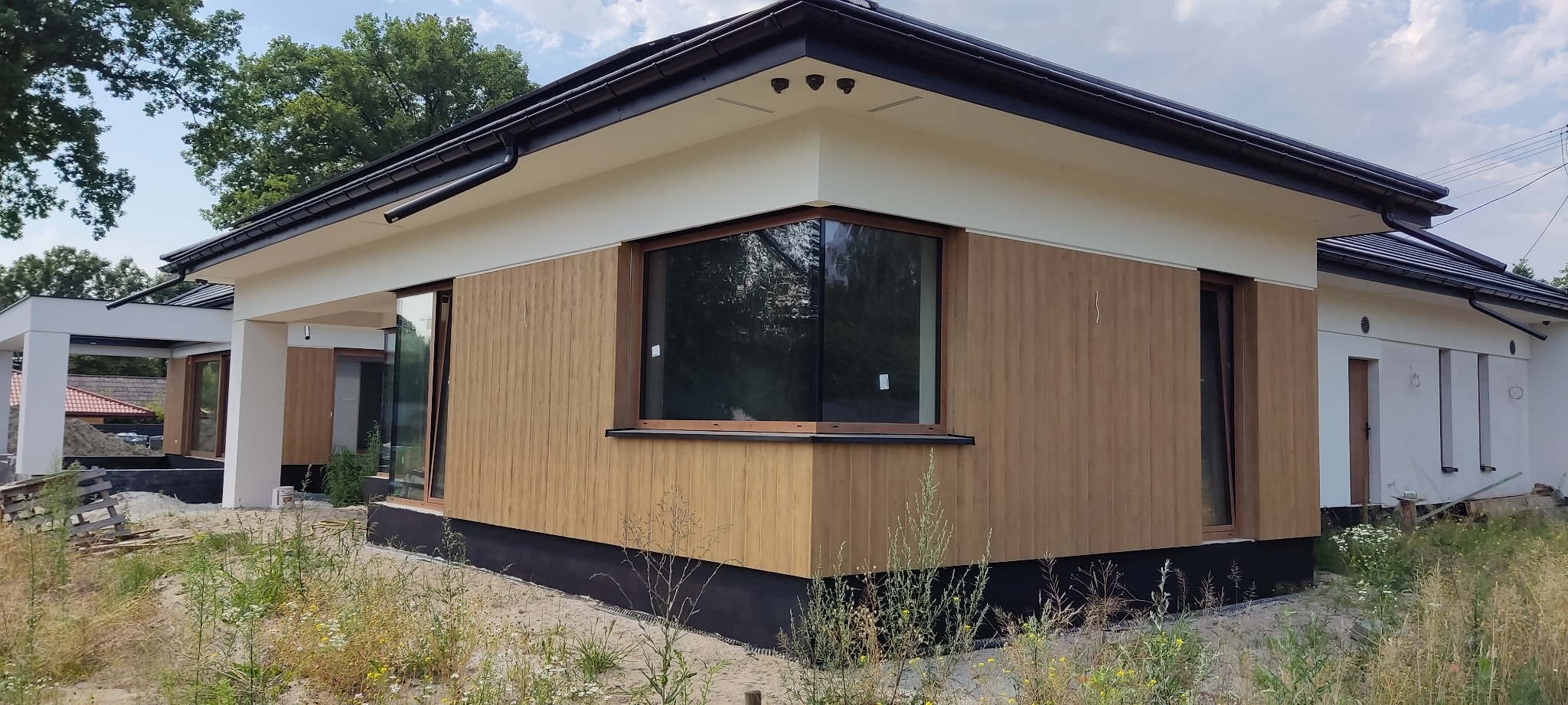 Nowoczesny dom jednorodzinny z elewacją w odcieniach brązu i beżu, widoczne okna z drewnianą ramą oraz czarna izolacja fundamentów, otoczony zielenią.