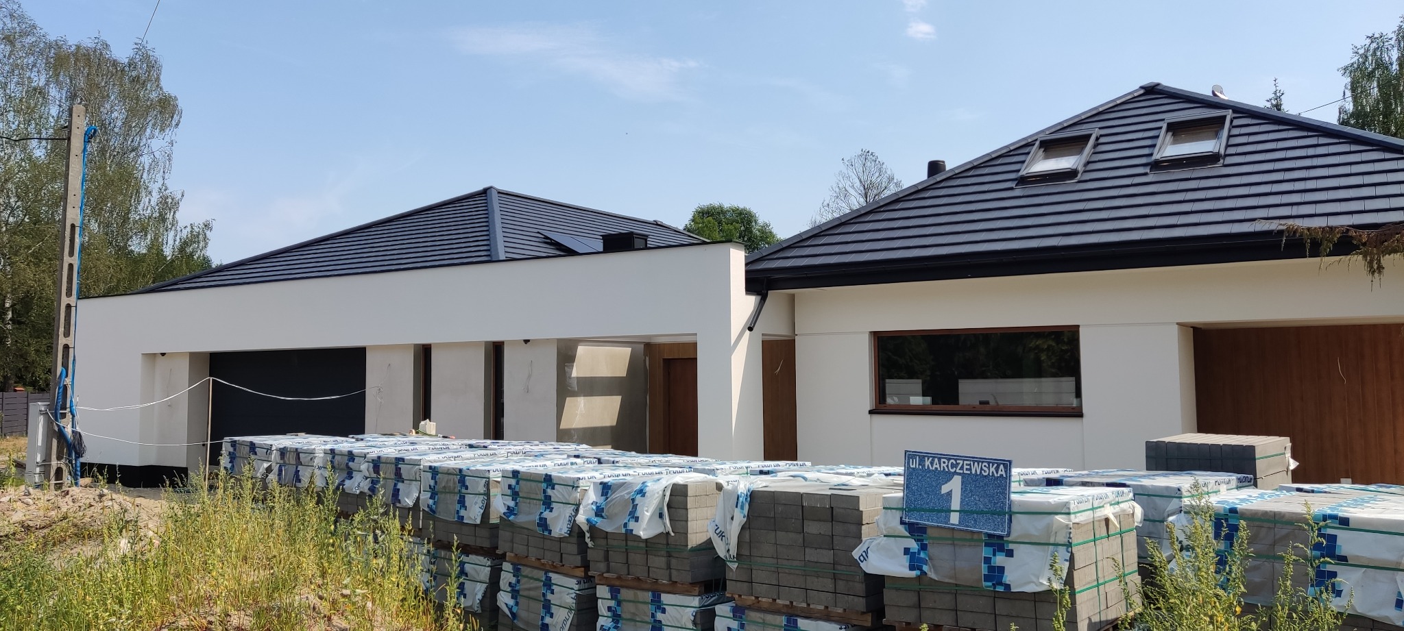 Nowoczesny dom jednorodzinny w trakcie budowy, z widocznymi paletami kostki brukowej na pierwszym planie i tabliczką z adresem 'ul. Karczewska 1'.