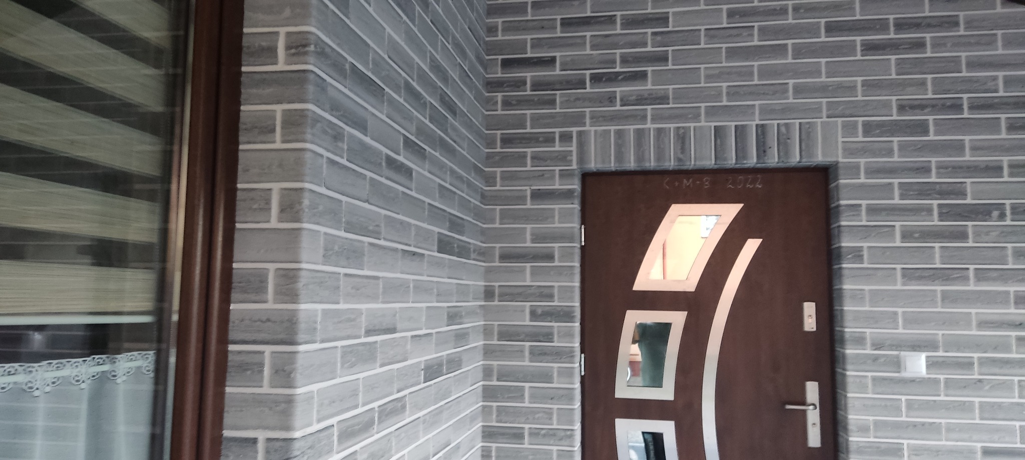 Elewacja budynku wykończona szarą cegłą klinkierową, brązowe drzwi wejściowe z geometrycznymi przeszkleniami, widok na okno z żaluzjami.