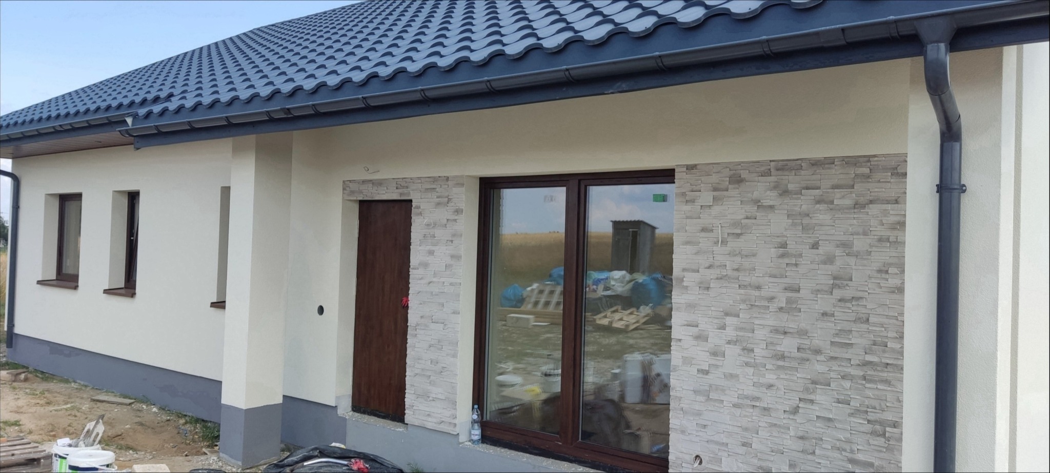 Wykończona elewacja domu jednorodzinnego z fragmentami dekoracyjnej okładziny imitującej kamień wokół okna i drzwi, widoczne rynny i fragment dachu z dachówki.