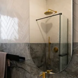 Nowoczesna łazienka z lustrem owalnym, ukazującym złotą armaturę prysznicową i umywalkową, oraz biało-szare płytki ułożone w jodełkę powyżej marmurowej ściany