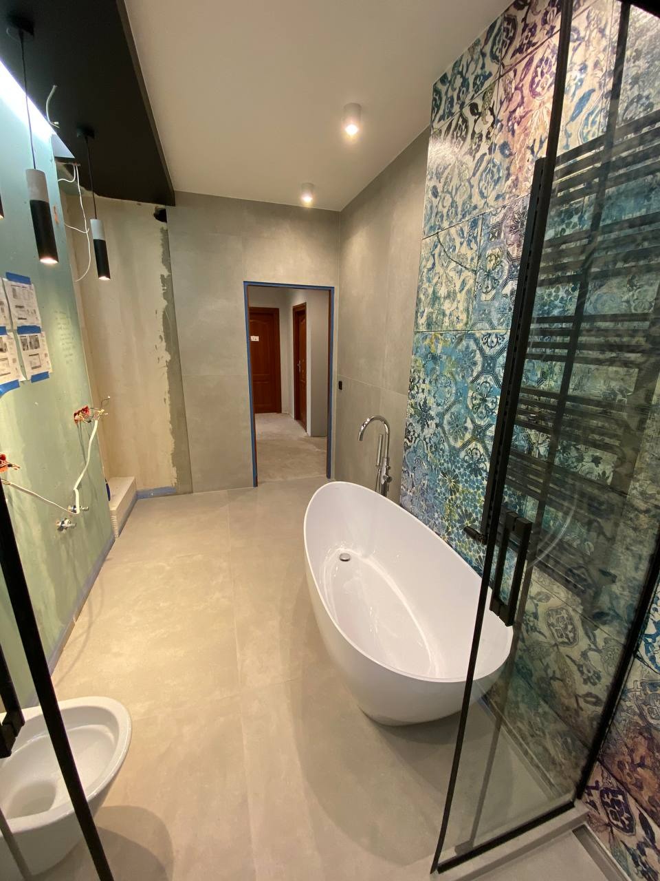 Wnętrze łazienki w trakcie prac wykończeniowych: owalna wanna wolnostojąca, fragment kabiny prysznicowej z czarnymi profilami, ściana z dekoracyjną, wzorzystą glazurą, surowa ściana z widocznymi...