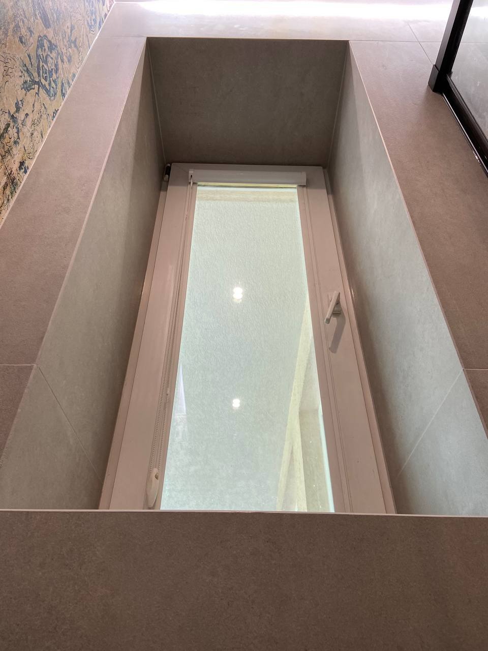 Wnęka okienna w łazience wyłożona płytkami w odcieniach szarości, z widoczną roletą okienną. Na ścianie po lewej stronie dekoracyjna tapeta lub płytki imitujące tapetę.